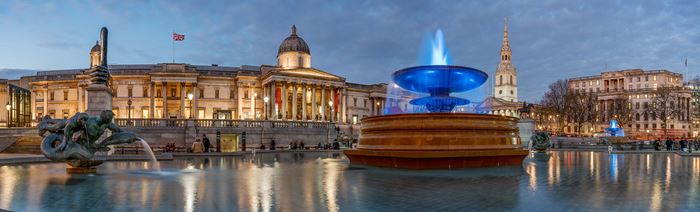 Trafalgar Square at sunset with fountains and buildings in the background lit up, including the National Gallery.