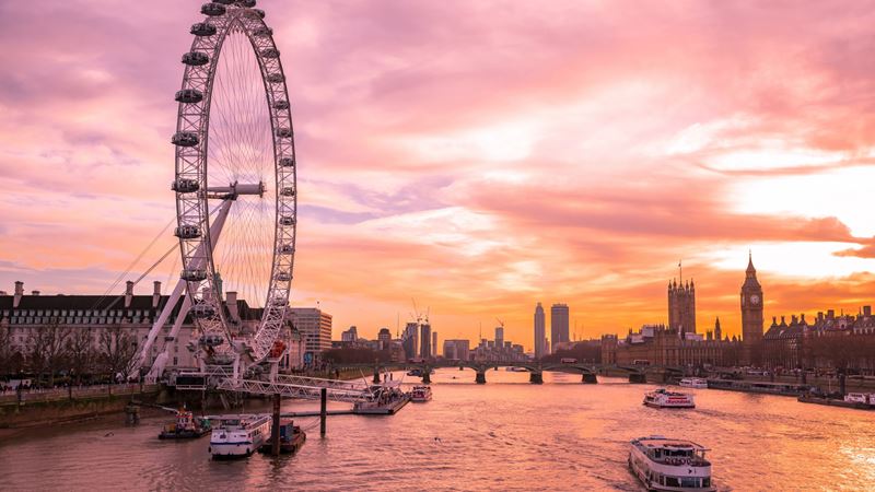 River Thames and London skyline at sunset with boats gliding along and Big Ben in the background.