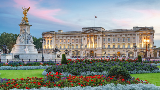 Buckingham Palace. Image courtesy visitlondon.com/Jon Reid. Red flowers growing in front of Buckingham Palace, which is lit up and sits in front of a blue and pink sky.