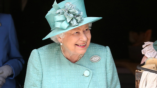 The Queen dressed in a turquoise jacket and hat, looking away from the camera and smiling.