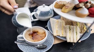 afternoon tea stand with sandwiches and sweet treats, and a person pouring milk into a cup of tea