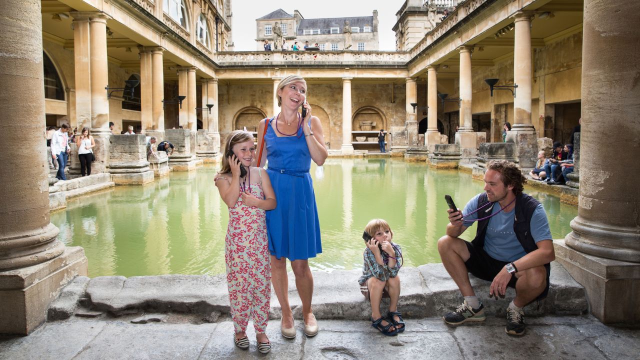 Uncover the history of the Roman Baths. Image courtesy of Golden Tours A family of four stand by the green waters of the roman baths in bath listening to the audio guide.