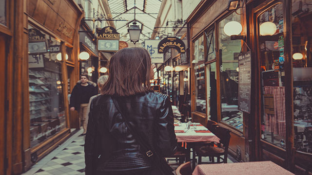 Passage des Panoramas in Paris. Photo by Tristan Colangelo on Unsplash A woman wearing a leather black jacket is walking across a wooden shopping gallery in Paris.