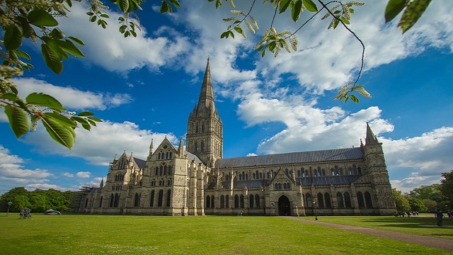 Landscape photo of Salisbury Cathedral, framed by green grass and shrubbery and blue cloudy skies in the background