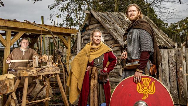 Discover life as a viking at the JORVIK Viking Centre in York. Image courtesy of Pyper York. Photo credit: Charlotte Graham. Two people dressed as vikings stand with a bright yellow and red shield and a man holds ancient tools in a brown workshop.