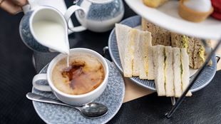 Milk is poured in a cup of black tea placed next to a plate of finger sandwiches.