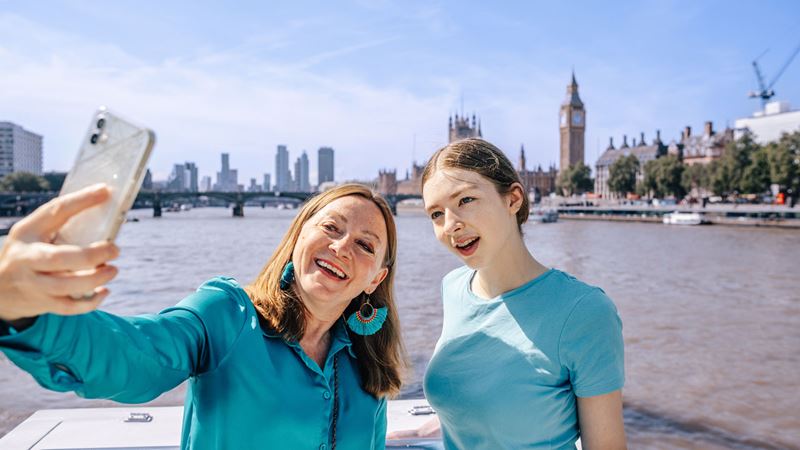 Explore London at your own pace using City Cruises sightseeing day pass. Image credit: Visit London/Michael Barrow. Two women are taking a selfie abord a City Cruises boat, with Big Ben on the background.