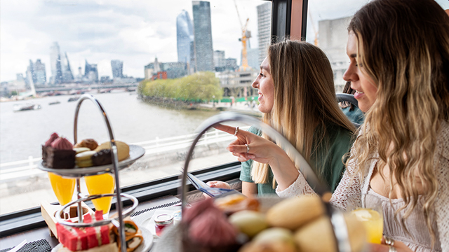 Two women are seated at a table with a set of cakes while lookin at the city views through the bus window.