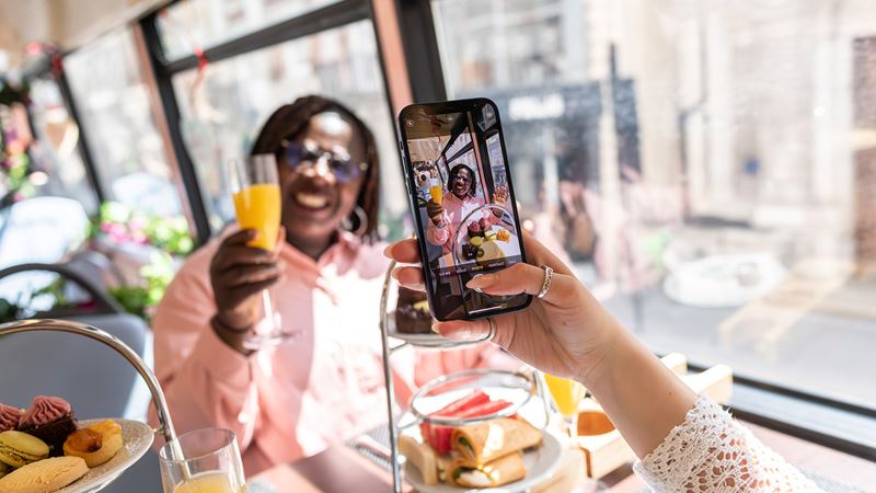 Take an afternoon tea aboard a panoramic tour of London, the perfect occasion for these famous selfies! Credit: London & Partners/Michael Barrow. Image courtesy of visitlondon.com. A woman is taking a photo on her smartphone of her friend holding a mimosa during the afternoon tea bus tour of London.