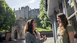 Two women discuss passionately while standing in the street with Windsor Castle shown in the backdrop.