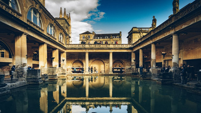 Go back in time with a visit to the Roman Baths in Bath. Photo by Paul Cuoco on Unsplash. View on the Roman Baths churchyard by a cloudy day with the building reflecting on the water.