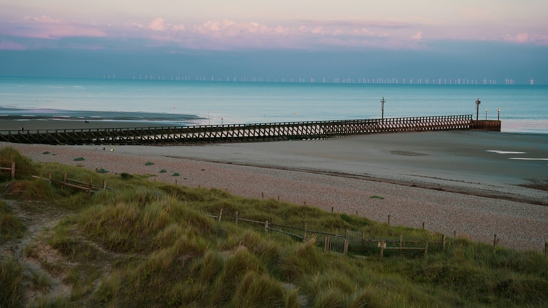 Littlehampton, West Sussex. Photo by Andy Gill on Unsplash. Deserted Littlehampton beach with grassy banks, shingle and a pontoon leading out to see at sundown.