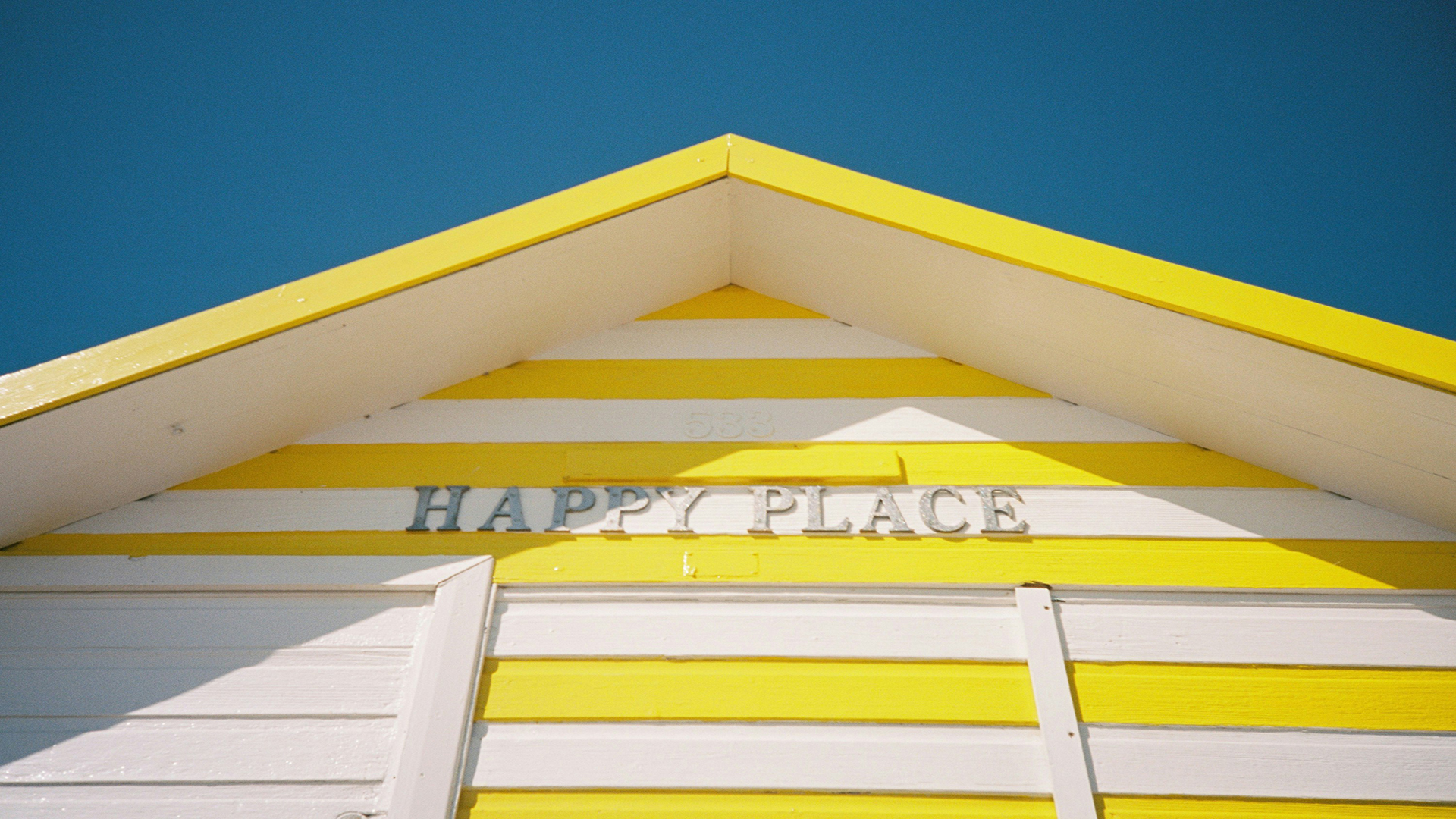 Walton-on-the-Naze, Essex. Photo by Nick Page on Unsplash. Close-up of a white and yellow stripe beach hut with the words 'happy place' and a clear blue sky in the background.
