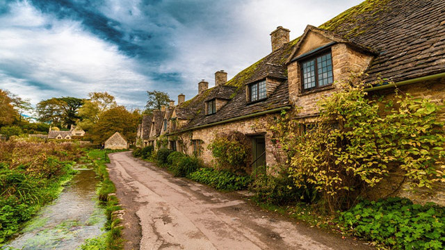Explore quaint villages in the Cotswolds. Photo by Samuel Sweet, Pexels. View of traditional stone houses alongside a path bordering a river in the countryside.