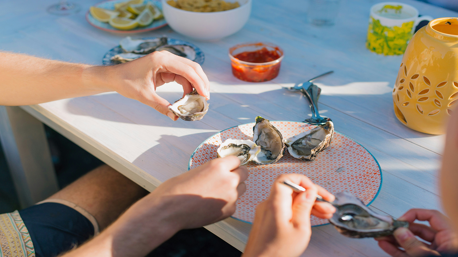 Tuck into delicious oysters in Whitstable. Photo by Charlotte Harrison on Unsplash. Close up of someone holding an oyster, with a plate of oysters and lemon slices on a wooden table on a sunny day in Whitstable.