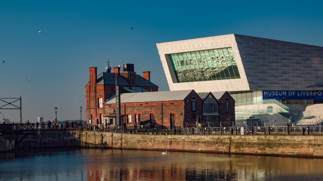 Museum of Liverpool buildings by the water on a clear day, with red-brick building and modern structure displaying Imagine Peace exhibition title in big letters on the front of the building. 