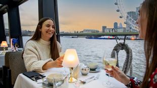 two women sat at a table on a cruise boat, cheersing with wine glasses and the sun setting in the background over the London Eye