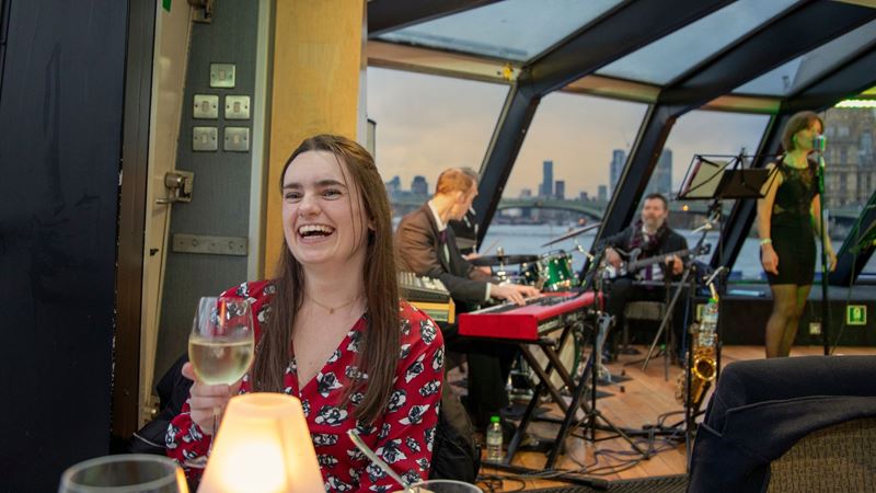 Savour a delicious three-course meal while cruising past iconic London landmarks, all set to the smooth sounds of live jazz. Image courtesy of Golden Tours A smiling lady, sat a table holding a glass of wine with a jazz band playing in the background