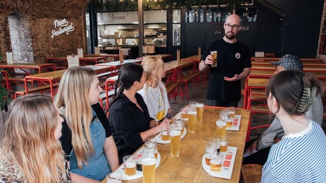 Learn all about Camden Town Brewery from an expert guide. Image courtesy of Golden Tours. A group of people sit at a long table trying different beers with a guide at a london brewery.