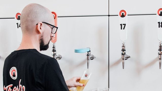 Try beers at Camden Town Brewery. Image courtesy of Golden Tours. A man wearing a black camden town brewery tshirt and glasses stands at beer taps pouring a pint of beer.