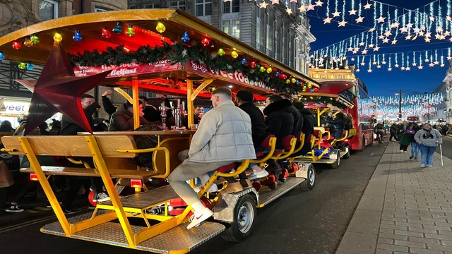 Hop aboard the Wee Toast Tour bike bar. Image courtesy of Wee Toast Tours. A group bike tour with 10 people on, cycling down a street in London with Christmas lights in the background.