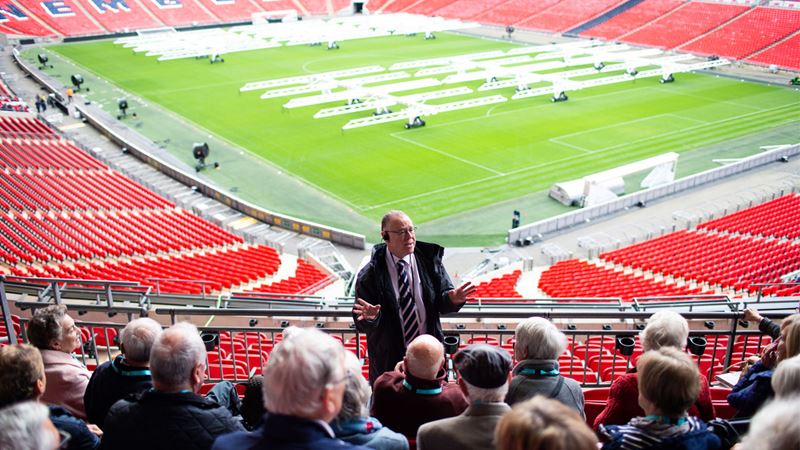 Guide talking in front of crowd of people in stands at Wembley Stadium with view of the pitch behind.
