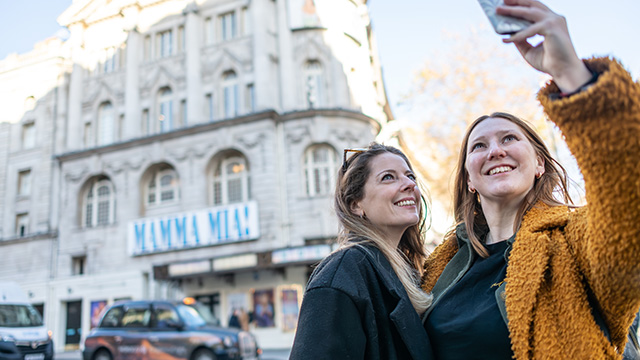 Two women take a selfie in front of a London theatre.