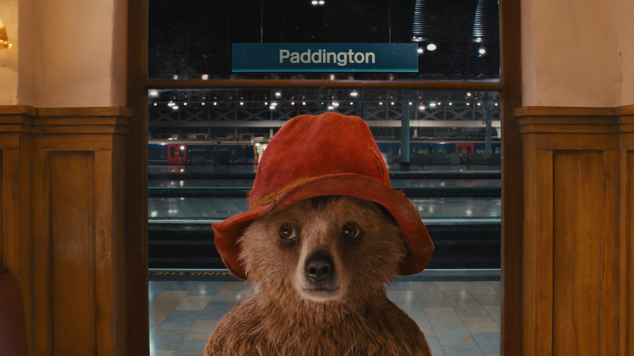 A scene from Paddington shot at Paddington Station. Paddington bear stands in Paddington train station wearing a red bucket hat.