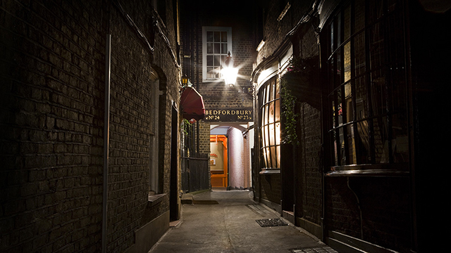 London Grim Reaper Tour. Image courtesy of Sandemans New London Tours. A narrow, darkened alley leads to a dimly lit pub at night, which is part of the London Grim Reaper Tour.
