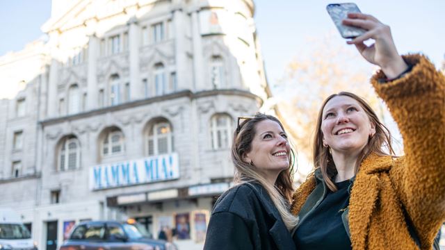 Two women taking a selfie in front of Mamma Mia theatre sign.