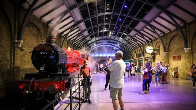 Board the Hogwarts Express! TM & © Warner Bros. Entertainment Inc. Harry Potter Publishing Rights © JKR. A tall man in grey takes a photo of a woman in a red jumper next to the red and black hogwarts express train at the warner bros studio tour.