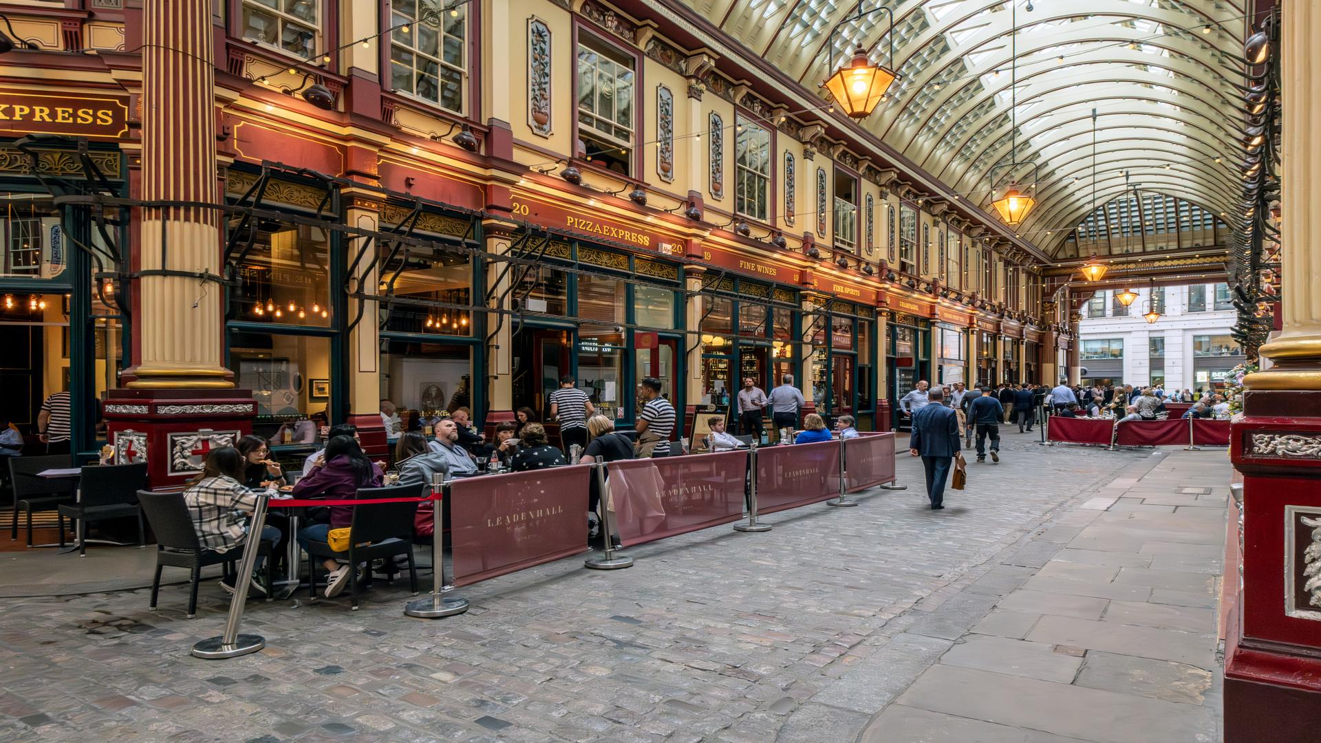Leadenhall Market, boasting a selection of boutiques, shops, restaurants and bars. Image courtesy of London & Partners / Michael Barrow. long angle shot of inside Leadenhall Market, with Victorian decor and people sitting outside a pub in the market