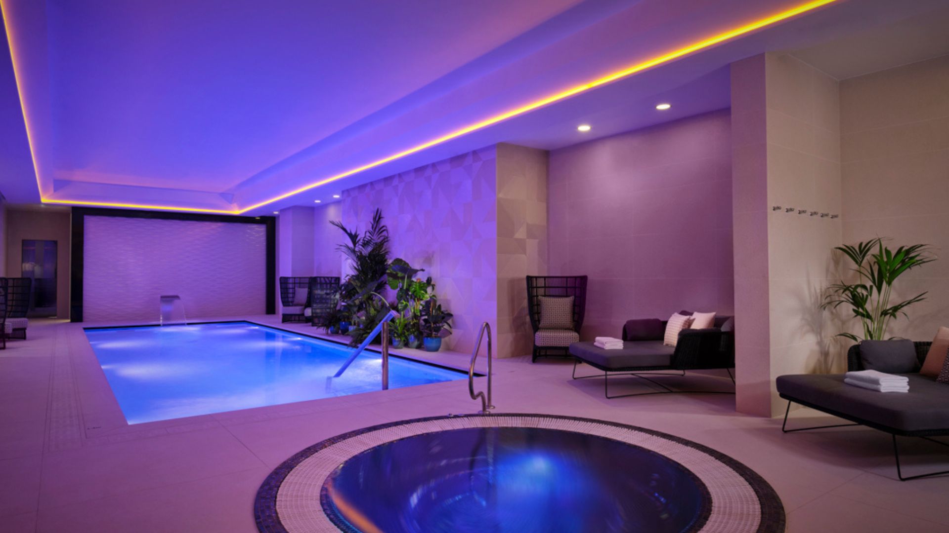 Book a romantic spa day at BOKEH by Montcalm East. Image courtesy of Red Letter Days. spa room with purple and blue mood lighting, a small round jacuzzi and a pool, with sofa seating on the side of the room