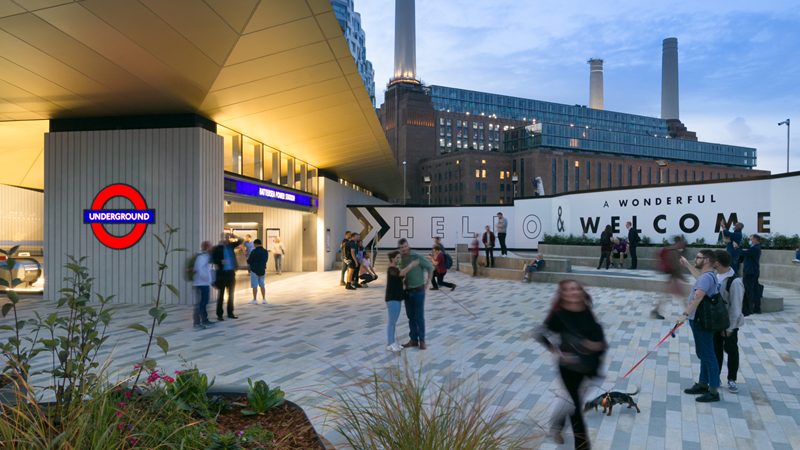 Battersea Power Station Tube station in the evening with people walking around in the foreground and the iconic Battersea Power Station with lights on in the background