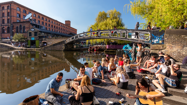 The sun shining down as people sit and eat next to Camden Lock and bridge