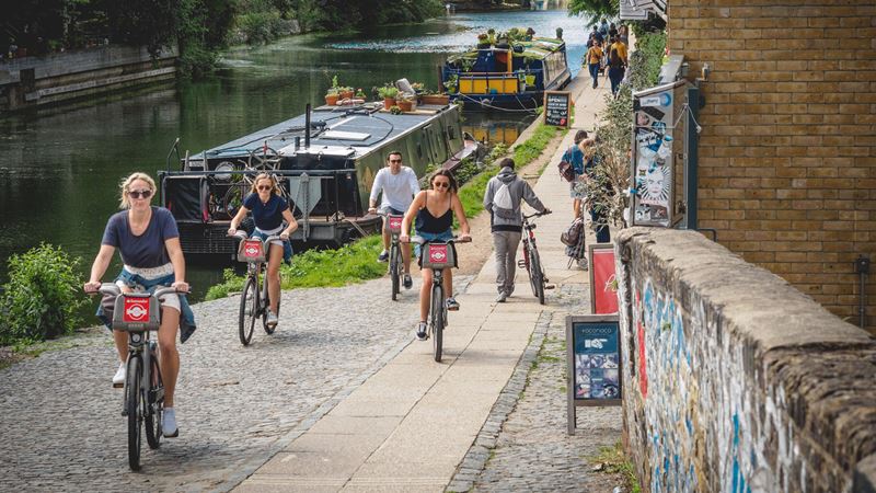 Tour the city by bike from a Santander Cycle. Credit: Shutterstock. Image courtesy of Shutterstock. A group of friends cycle next to the City Basin canal on Santander bikes with boats on the river in the background