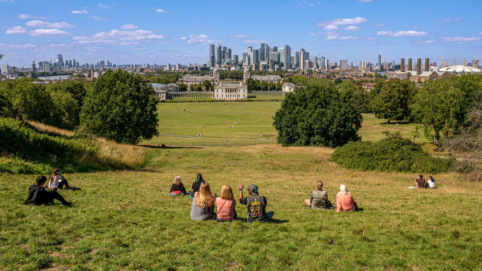 Groups of people sat on the grassy hill in Greenwich Park with a view of the National Maritime Museum and city in the background
