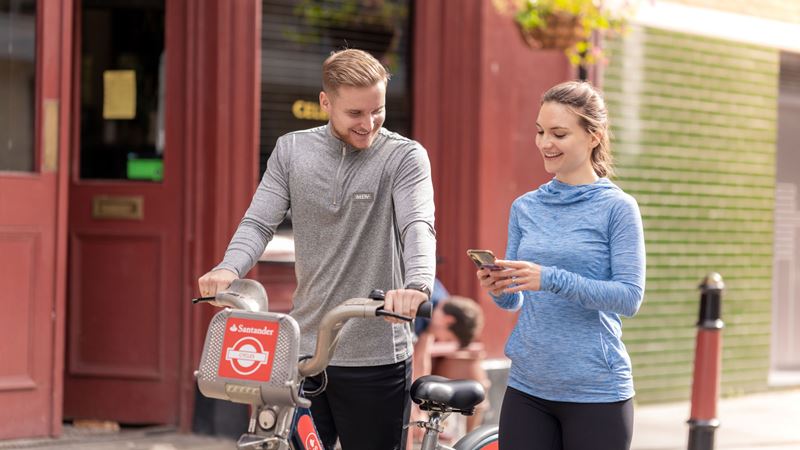 Have fun navigating the city using a Santander bicycle. Credit: visitlondon.com/Antoine Buchet. Image courtesy of Antoine Buchet. A man and woman walk along a street smiling at each other while the woman is on her phone and the man pushes a Santander bicycle