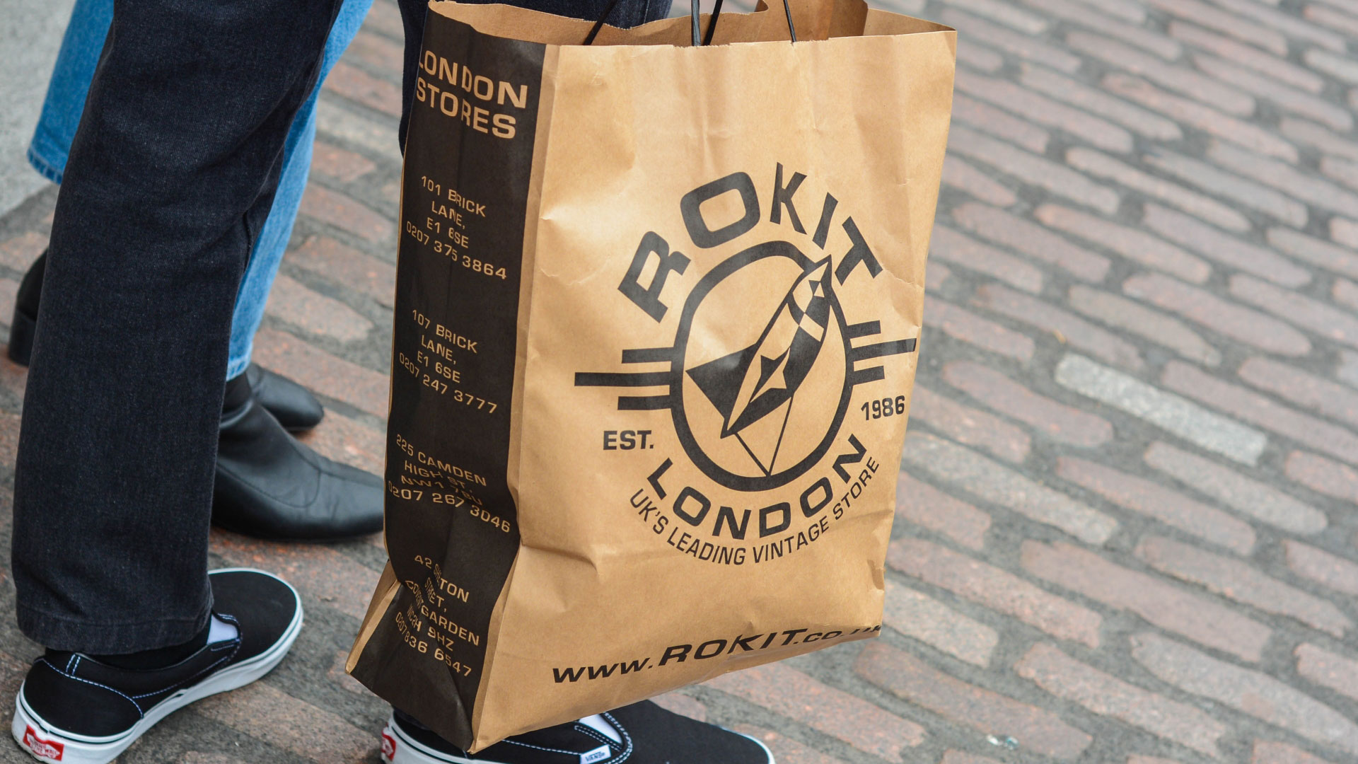 Browse the shelves of vintage gems at Rokit stores in London. Credit: Shutterstock. Image courtesy of Shutterstock. Someone holding a brown paper bag with the Rokit London logo on the front wearing jeans and black vans shoes