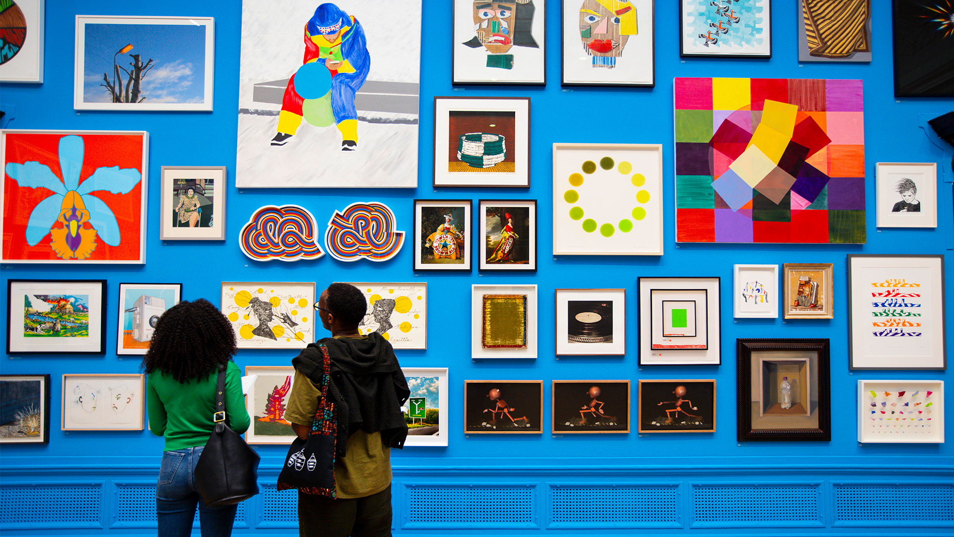 Installation view of the Summer Exhibition 2024 at the Royal Academy of Arts. Image courtesy of Royal Academy of Arts, London/David Parry. A couple looking at a wall covered in different artworks at the Summer Exhibition