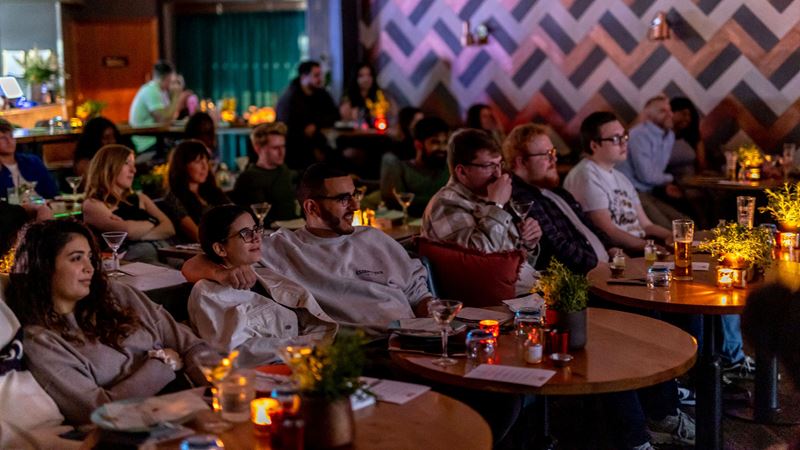 Embark on an immersive film journey during a Taste Film x Drake & Morgan dining experience. Credit: Victor Frankowski. Image courtesy of Taste FIlm. An audience of people sit in comfy chairs looking forward at a film screen with tables in front of them with lamps and drinks ready to be served food