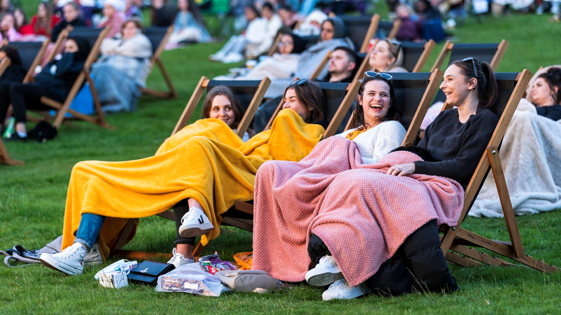 Watch a film at one of London's top outdoor cinemas. Image courtesy of Adventure Cinema. Four girls sit on deck chairs on grass in a park surrounded by other revellers with blankets over them ready to watch a film at an outdoor cinema screen