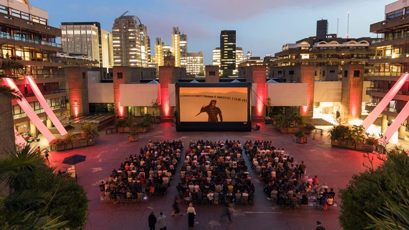 A photo from above of the Barbican Centre sculpture court with and audience of people facing a big screen playing Dune with red lights surrounding