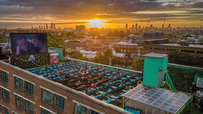 Make London's sunset the backdrop to your next movie date at Rooftop Film Club next in Peckham's Bussey Building. Credit: Rooftop Film Club. Image courtesy of Rooftop Film Club. A photo from above Rooftop Film Club in Peckham with the screen playing a movie to seats of people on the rooftop as the sun sets behind London's buildings in the distance