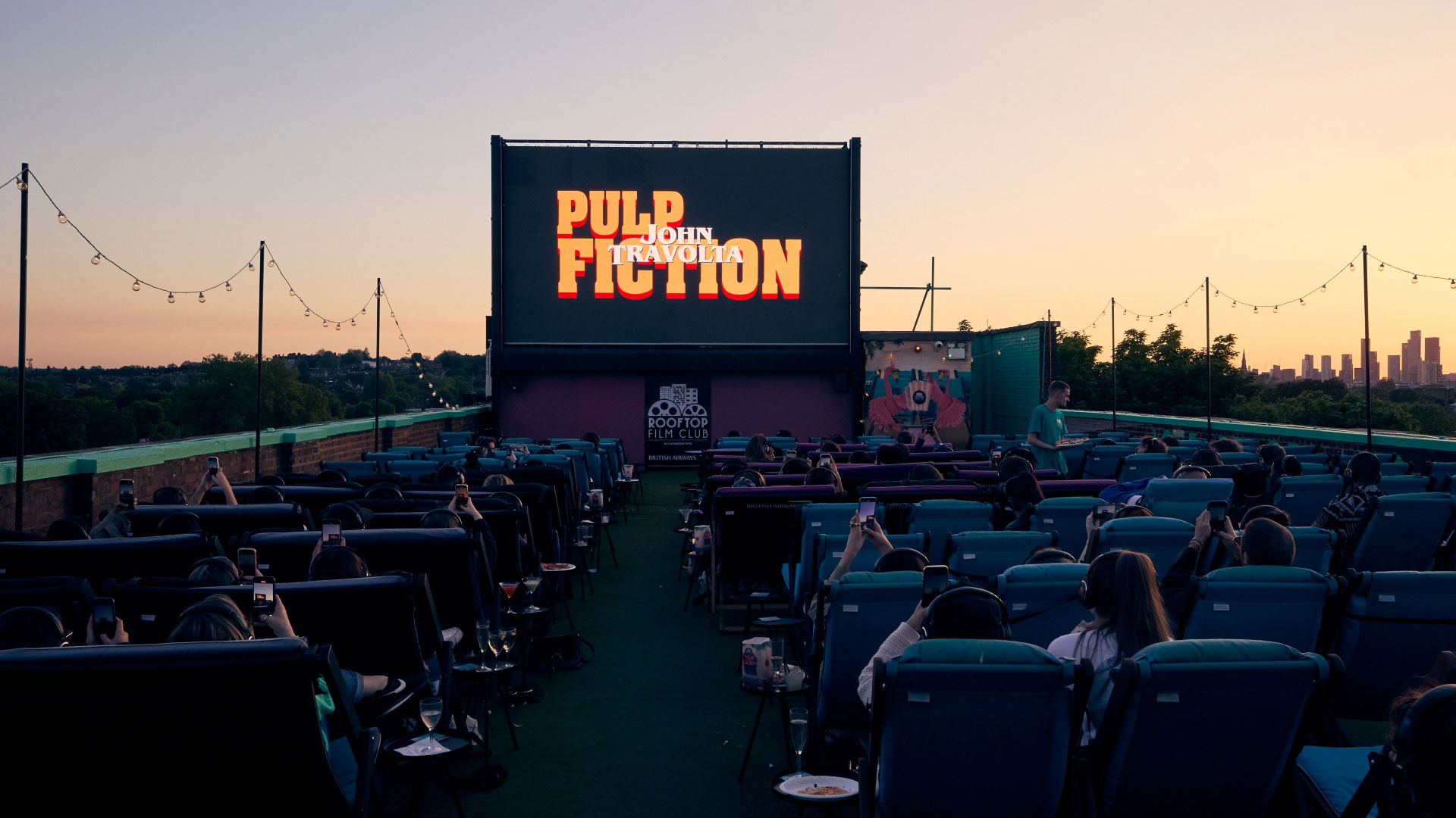 Catch a film beneath the stars at Rooftop Film Club this weekend. Image courtesy of Rooftop Film Club. A photo of Rooftop Film Club Peckham from behind the seats of people with the screen showing "Pulp Fiction" title screen as the sun sets in the background