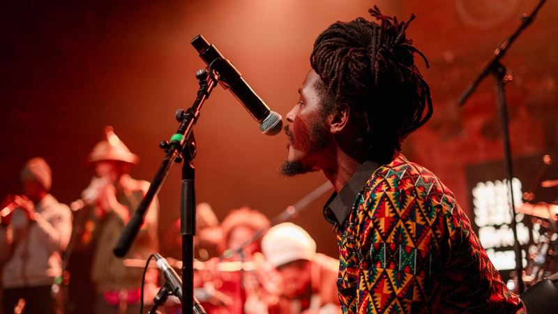 Listen to London's best underground music at the Brick Lane Jazz Festival. Credit: Ilhan Beyazay. Image courtesy of Prescription PR. A black musician singing live with his band in the background and dim red lighting at the Brick Lane Jazz Festival.