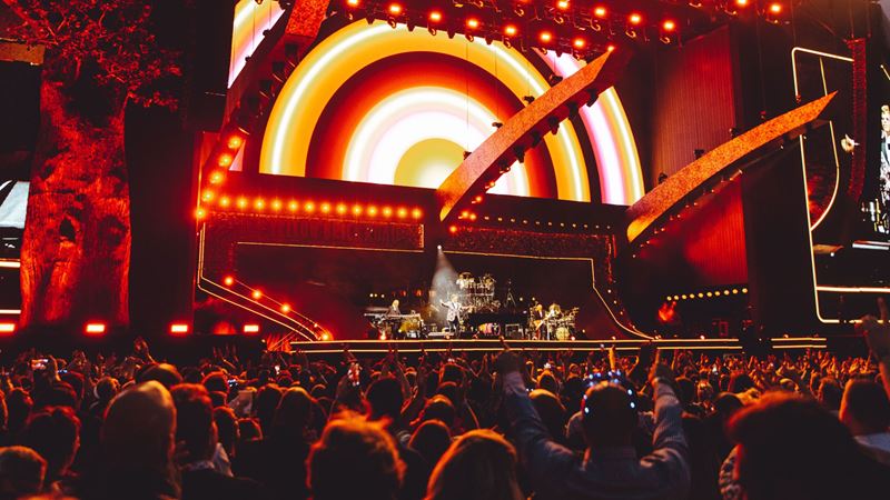 Join the crowds of fans that flock to Hyde Park for its annual summer festival. Credit: Hyde Park British Summer Time. Image courtesy of Hyde Park British Summer Time. A photo from the crowd of the stage at Hyde Park British Summertime with bright orange coloured lights and visuals on the screens forming the backdrop of the stage