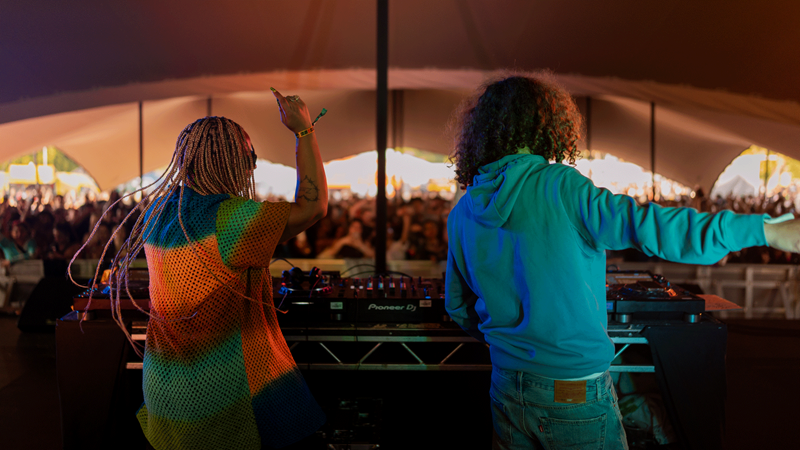 Behind the DJ decks at City Splash. Credit: Eljay Briss. A girl in a rainbow shirt and man in a blue hoodie DJ in a tent in front of large audience