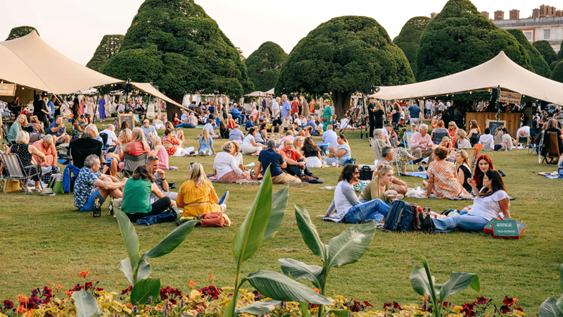 Unpack your picnic and pop a bottle of fizz in the gardens during Hampton Court Palace Festival. Credit: Joshua Atkins. Image courtesy of Joshua Atkins. Crowds of festival goers sit in groups under the sun enjoying picnics on the grass