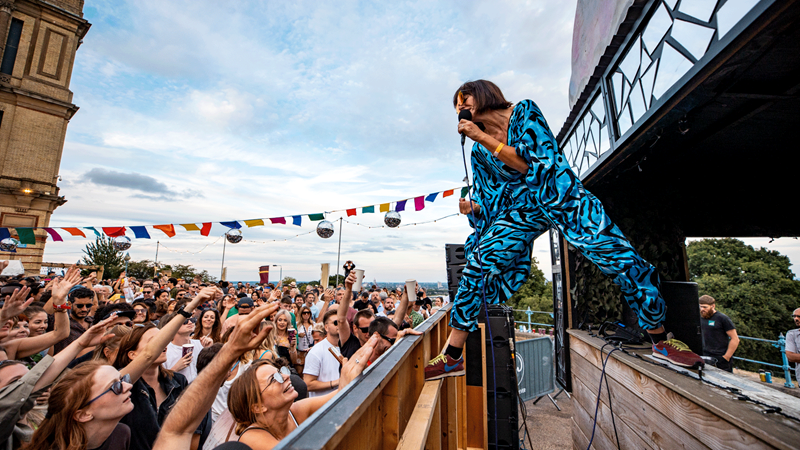 Spend the day dancing in the sunshine at the Kaleidoscope Festival Outdoor Stage. Credit: Lloyd Winters. Image courtesy of Lloyd Winters. Performer in a blue jumpsuit sings to the crowd looking out across Alexandra Palace.